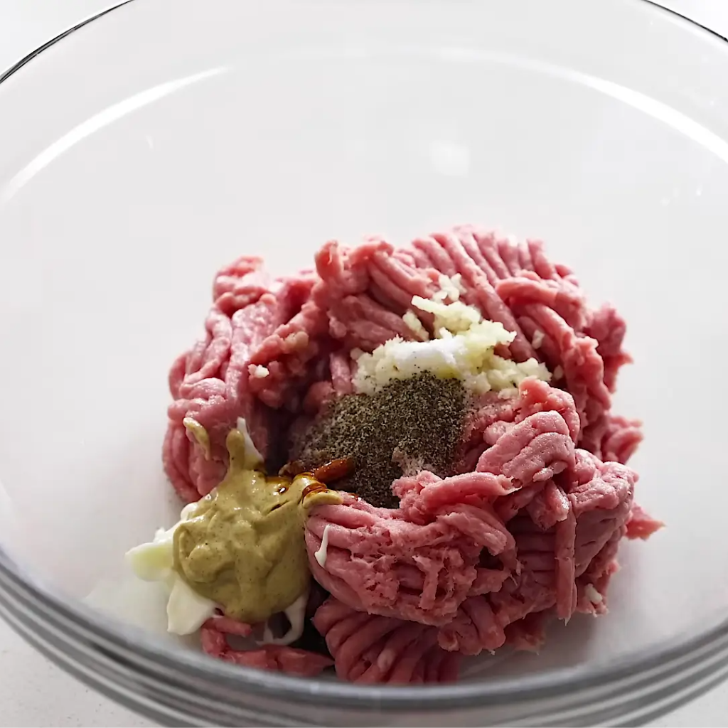 Raw ground meat in a clear glass mixing bowl topped with minced garlic, salt, pepper, Dijon mustard, mayonnaise, and Worcestershire sauce, ready for mixing into burger patties.