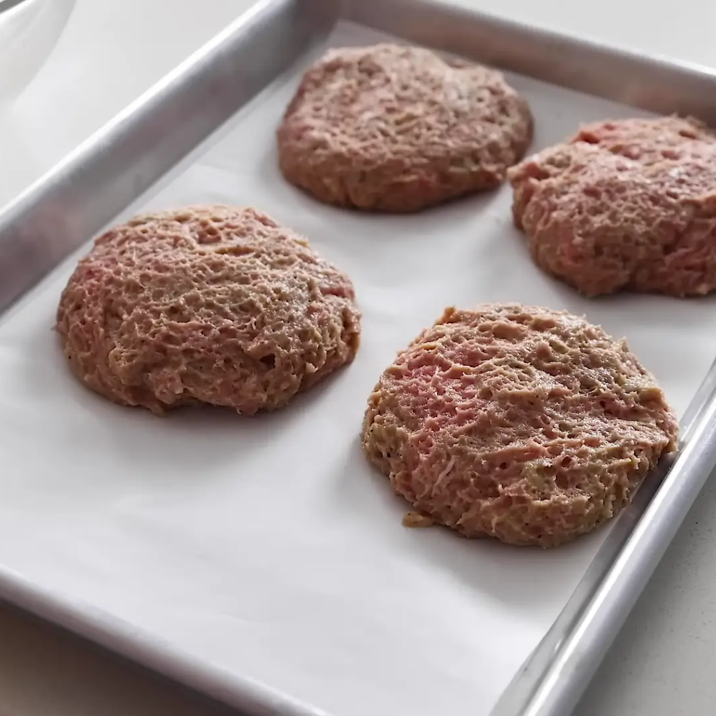 Four uncooked burger patties formed and arranged on white parchment paper lining a metal baking sheet, ready for cooking.