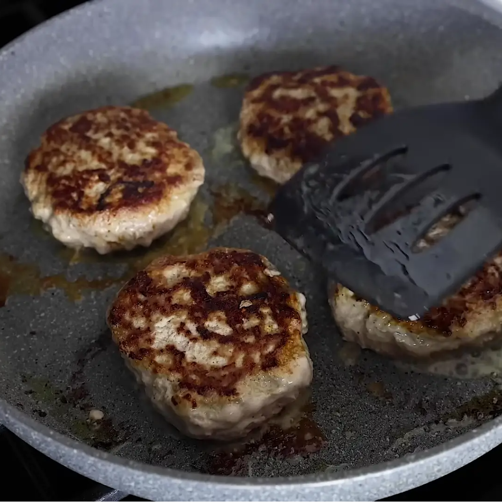 Four turkey burger patties cooking in a grey skillet, browned on one side, with a black spatula flipping one patty over.