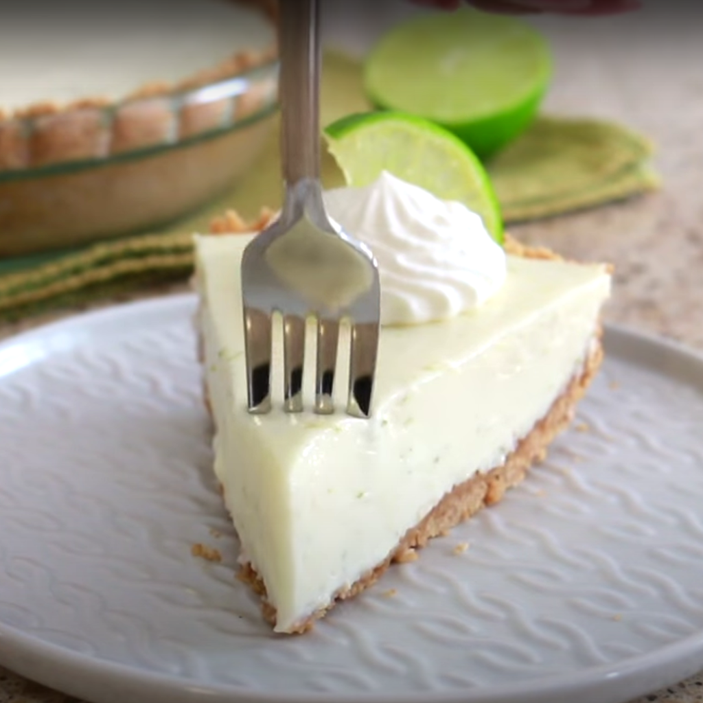 "A fork gently presses into a smooth, creamy slice of no-bake key lime pie, topped with a swirl of whipped cream, served on a white patterned plate. Halved limes and the rest of the pie are blurred in the background.