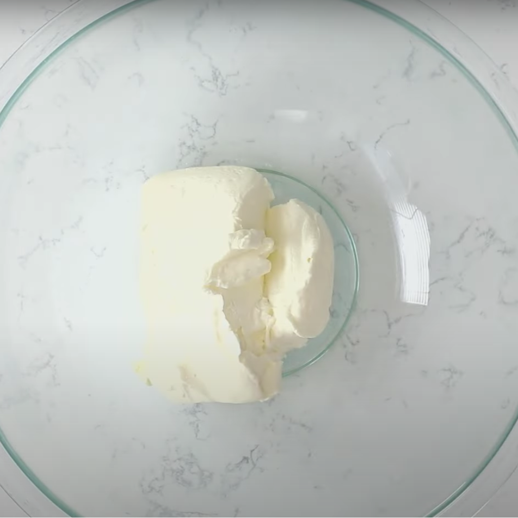 Top-down view of a block of white cream cheese in a clear glass mixing bowl on a marbled surface