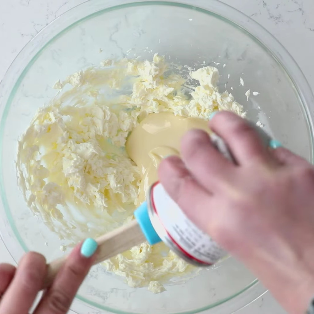 Hands pouring sweetened condensed milk from a can into a large glass mixing bowl containing a fluffy white ingredient, likely whipped topping, to make the filling for a no-bake key lime pie