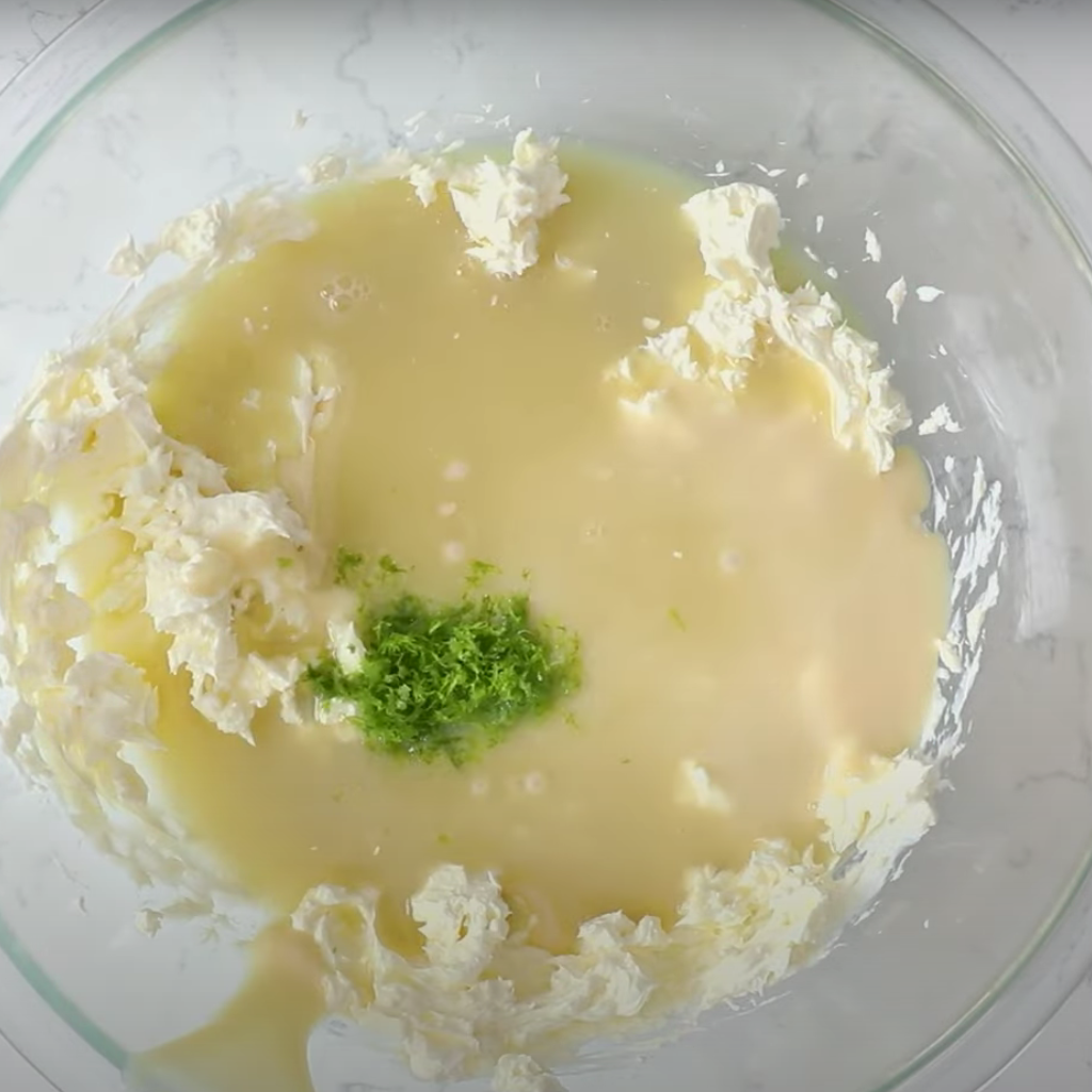 A glass mixing bowl seen from above, holding the ingredients for no-bake key lime pie filling: whipped topping, sweetened condensed milk, lime juice, and a vibrant pile of fresh lime zest in the center.