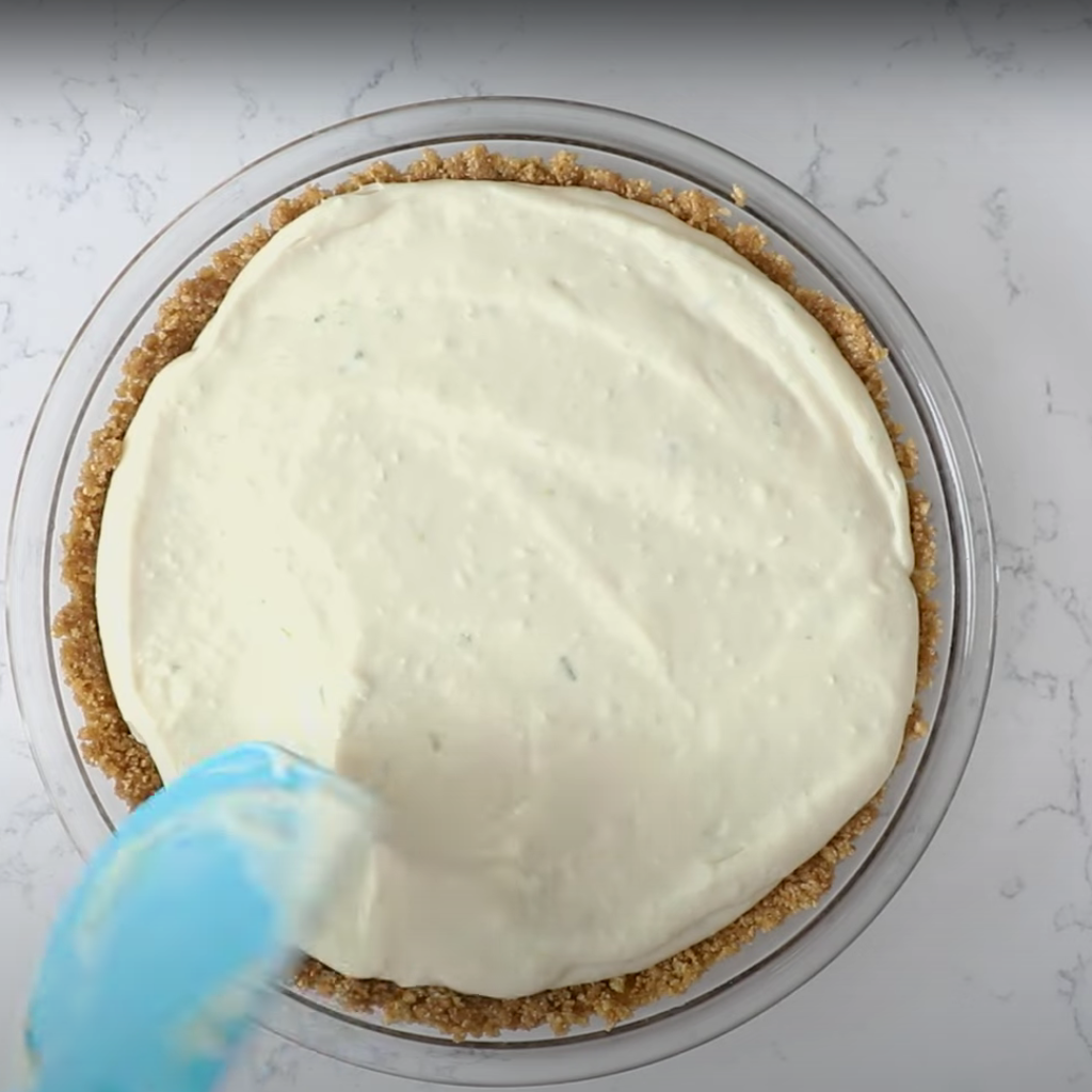 A blue spatula spreads creamy, pale green no-bake key lime pie filling smoothly into a golden graham cracker crust within a clear glass pie dish, seen from above.
